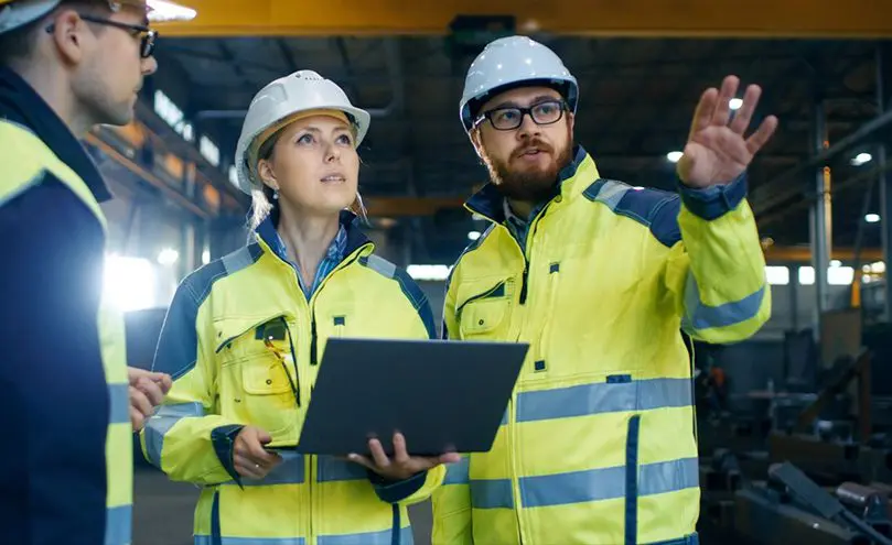 Two people in yellow jackets and hard hats holding a laptop.